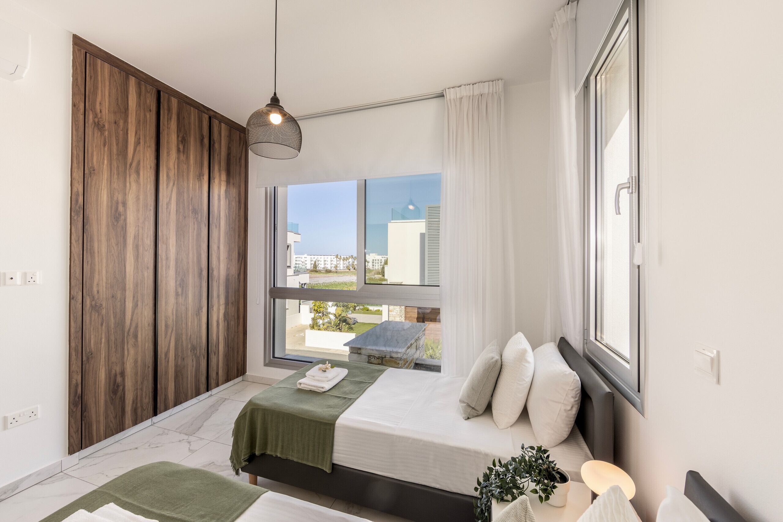Ein Schlafzimmer mit großem Fenster, einem Bett mit grüner Decke, einem Holzschrank und einer Hängeleuchte.