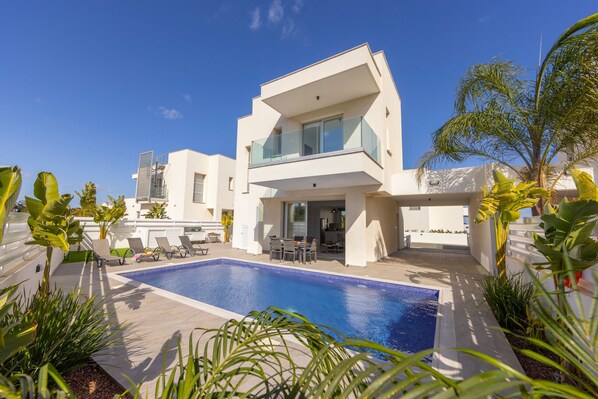 Une maison moderne à deux étages, dotée d’une piscine, d’un espace salon extérieur et d’une végétation luxuriante.