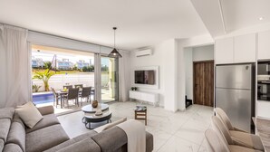 A modern living room with a sofa, TV, and a dining area with a pool view. - Protaras Sol e Mar Villa 2 (Paralimni)