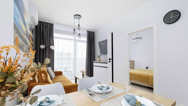 A modern dining area with a wooden table set for two, a sofa, and a TV in the background. - Seaside Apartment | 300 m to the Beach, Pool, Parking (Kołobrzeg)