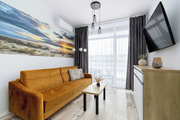 A living room with a mustard-colored sofa facing a wall-mounted TV. A small coffee table stands in front of the couch. Large windows with dark curtains brighten the interior.