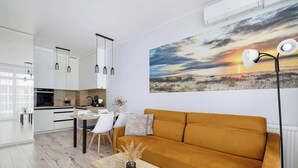 A modern kitchen with white cabinets, a dining area with a table and chairs, and a living room with a sofa and a painting of a sunset. - Seaside Apartment | 300 m to the Beach, Pool, Parking (Kołobrzeg)