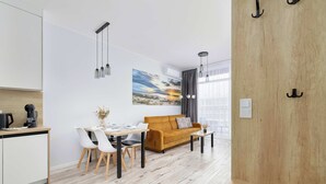 A modern living room with a wooden floor, a sofa, a dining table with chairs, and a kitchen area with a coffee machine. - Seaside Apartment | 300 m to the Beach, Pool, Parking (Kołobrzeg)
