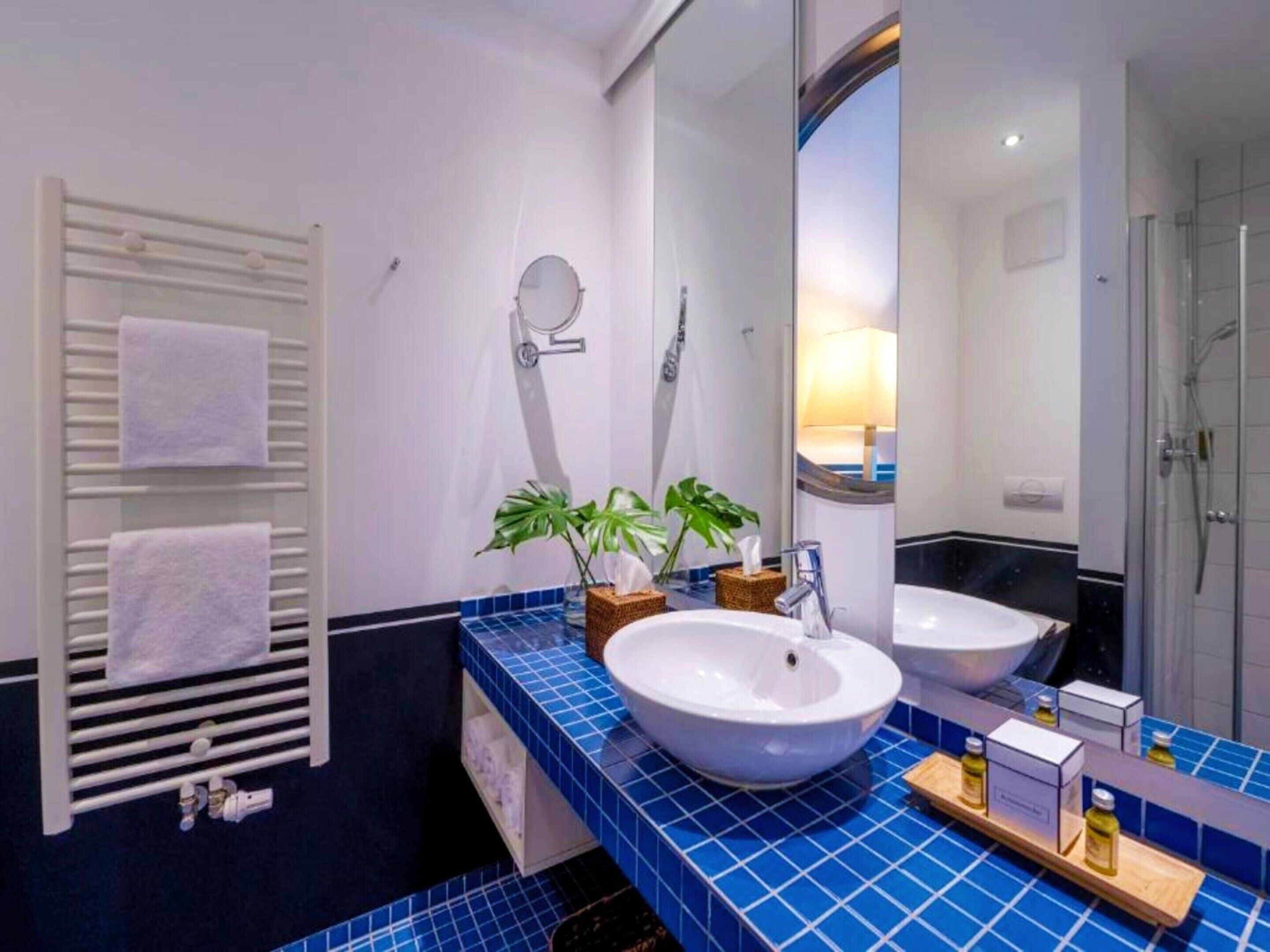 Ein Badezimmer mit einem weißen Waschbecken, zwei Spiegeln, einem Handtuchhalter mit weißen Handtüchern, einer Topfpflanze und einer Duschkabine.