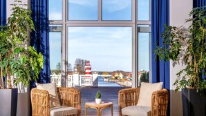 A room with two wicker chairs, a small round table, and large windows offering a view of a lighthouse and buildings across a body of water. - Sunday Resort Hafendorf Junior Suite (Rheinsberg)