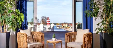 Una habitación con dos sillas de mimbre, una pequeña mesa redonda y grandes ventanales que ofrecen vistas a un faro y edificios al otro lado del agua.