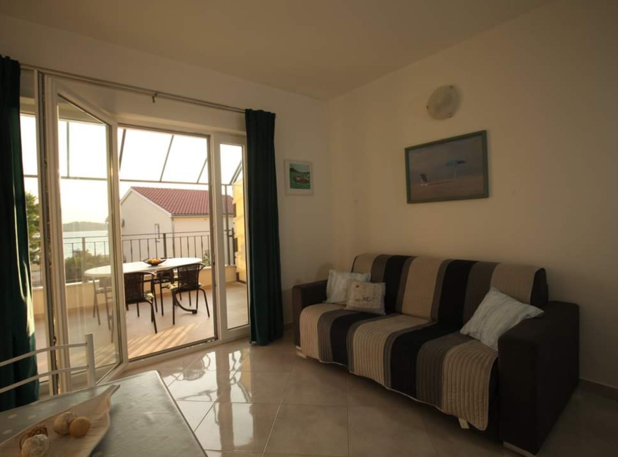 A living room with a sofa, a dining table with chairs, a sliding glass door leading to an outdoor area with a table and chairs, and a painting on the wall.