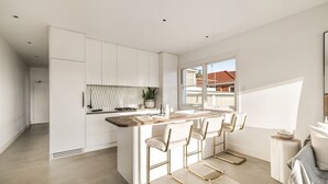 A modern kitchen with white cabinetry, a wooden countertop, and a large window. There are three bar stools with gold frames and a small island with a sink and stools. - Sunny Side Up North Bondi (North Bondi)