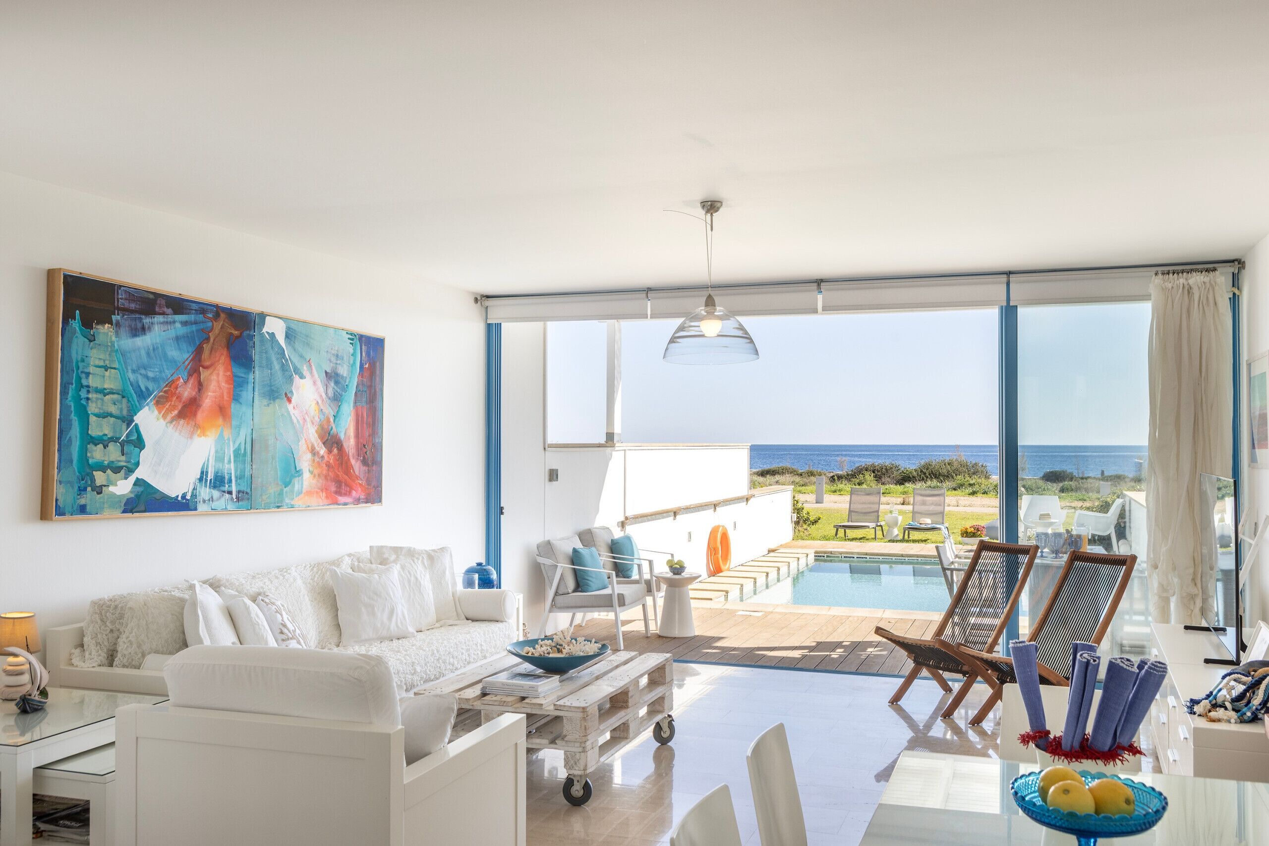 A modern living room with a large painting, a white sofa, a coffee table, and a view of the ocean through large windows.