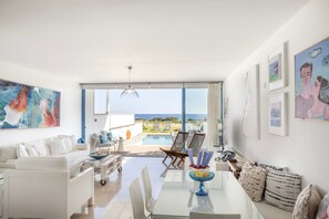 A spacious living area with a white sofa, a dining table with chairs, and a view of the ocean through large windows. - Protaras Seafront Villa Galene Del Mar (Paralimni)