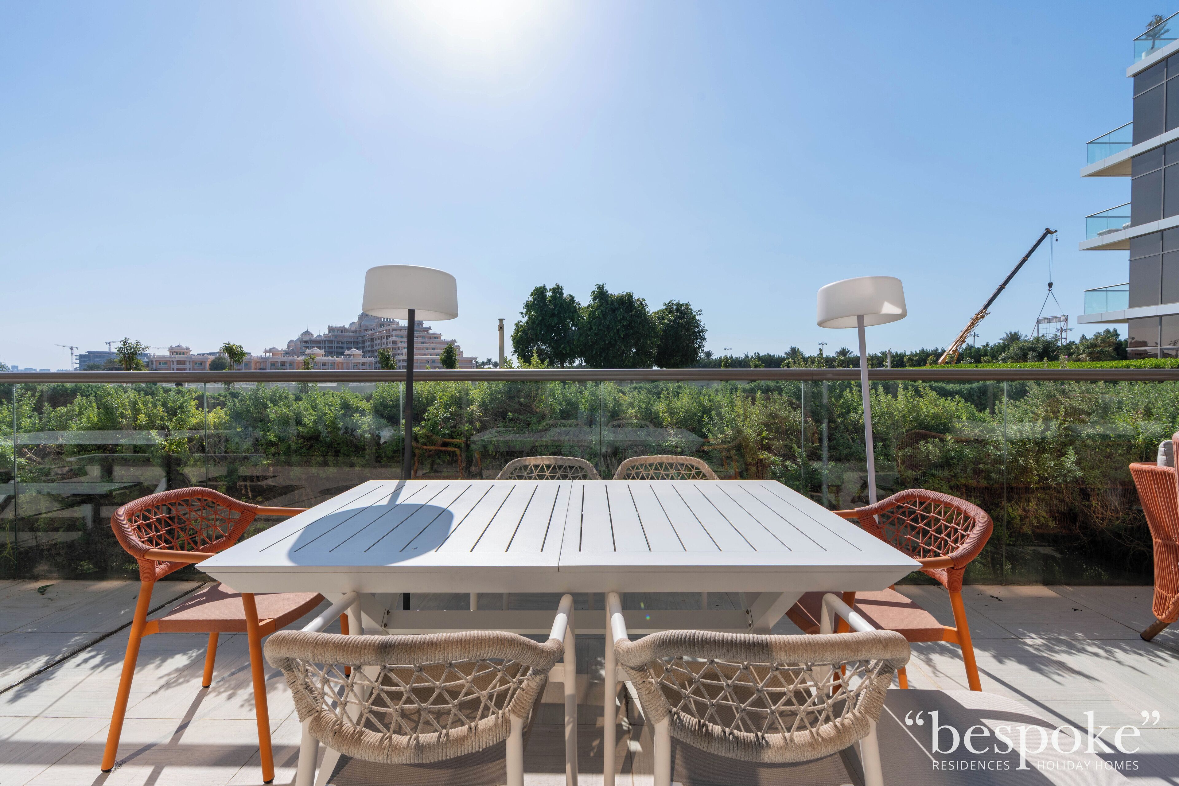 An outdoor dining area with a white table and wicker chairs, overlooking a landscaped garden and a construction site in the background.