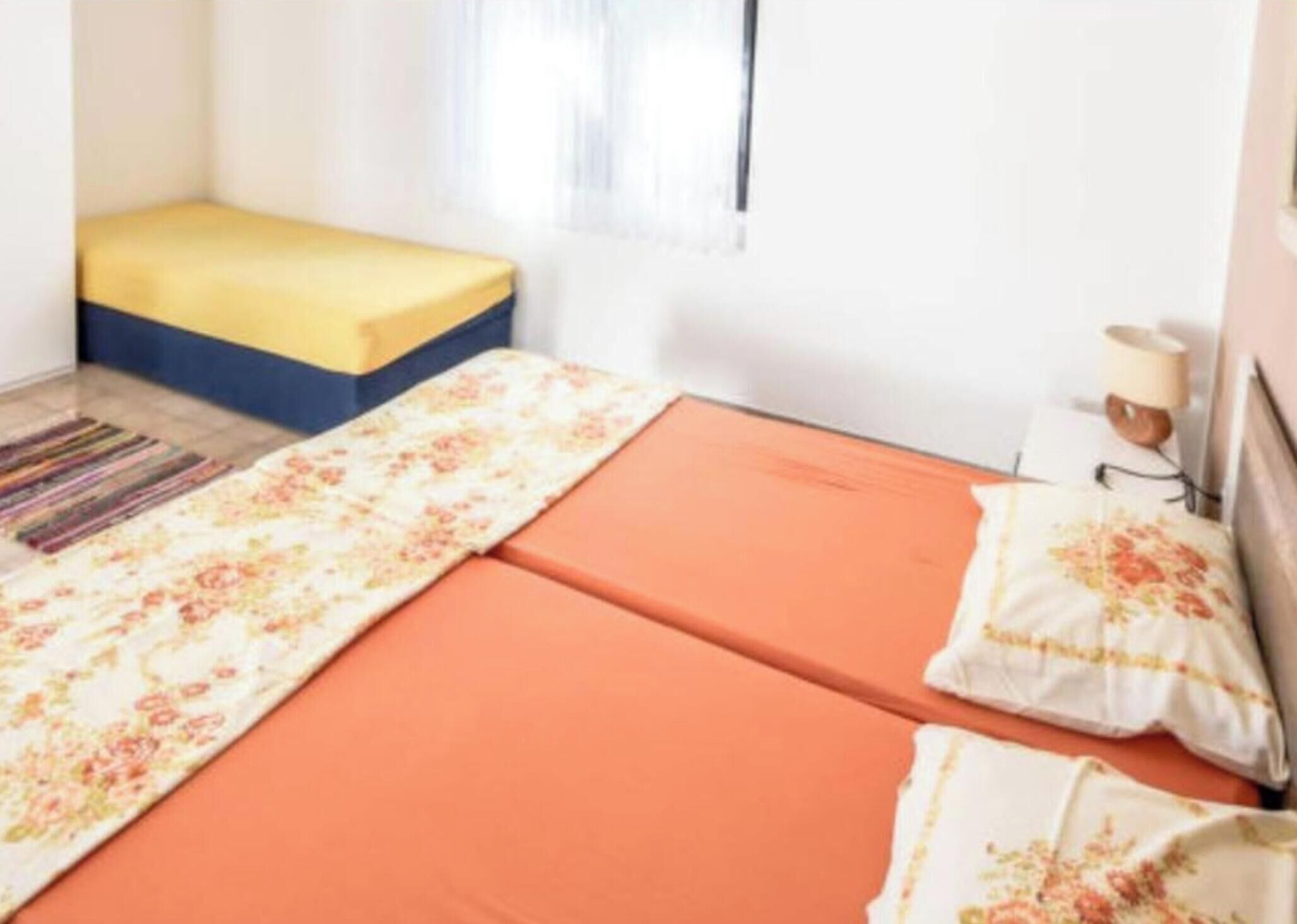 Une chambre avec deux lits, l’un avec un matelas jaune et l’autre avec un matelas orange. Il y a une fenêtre avec des rideaux blancs, une table de chevet avec une lampe et un tapis rayé sur le sol.