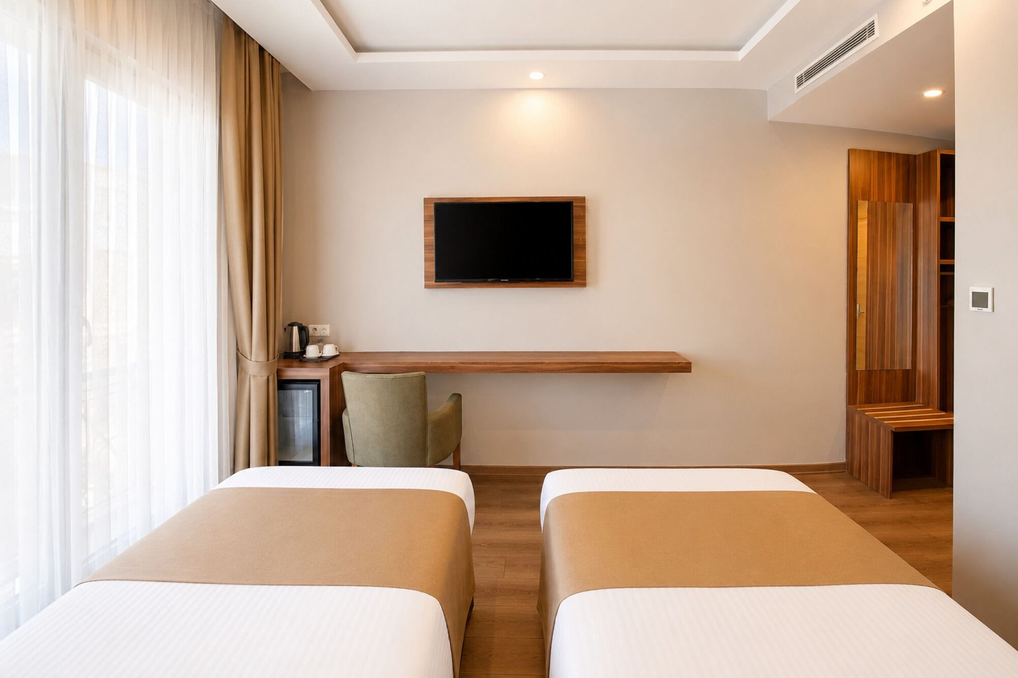 Quarto de hotel com duas camas, uma televisão de tela plana na parede, uma mesinha com chaleira e xícaras, e uma janela com cortinas leves.