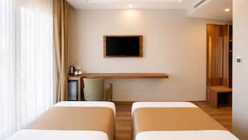 Quarto de hotel com duas camas, uma televisão de tela plana na parede, uma mesinha com chaleira e xícaras, e uma janela com cortinas leves.