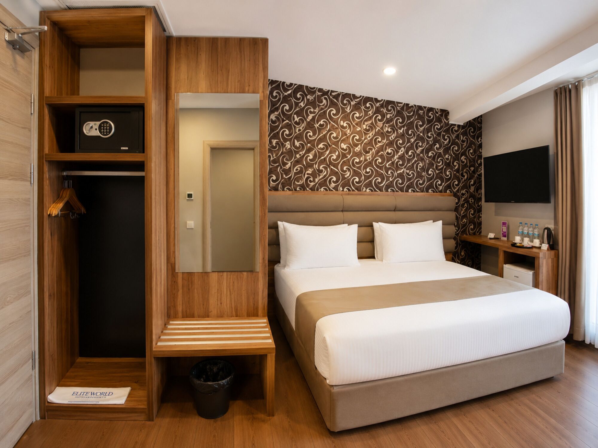 Ein Hotelzimmer mit einem großen Bett, einem Schrank inklusive Safe, einem Spiegel, einem Nachttisch, einem Fernseher und einem kleinen Schreibtisch mit Kaffeemaschine.