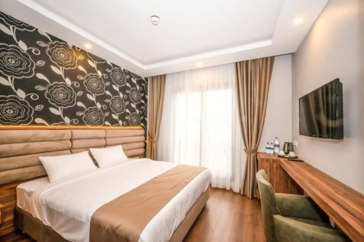 A hotel room with a large bed, a headboard with a floral pattern, a wooden desk, and a television on the wall.