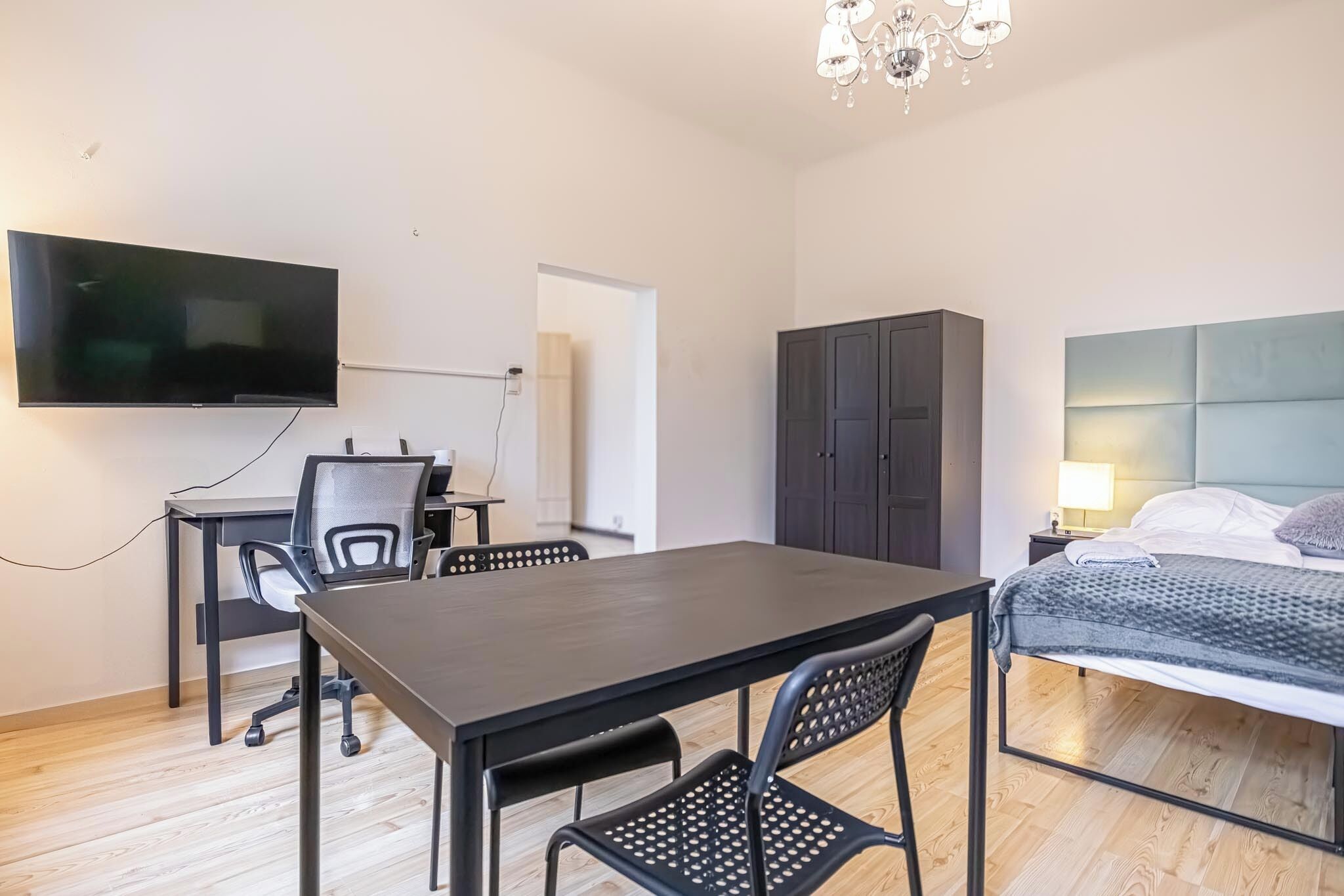 A modern hotel room with a flat-screen TV, a dining table with chairs, a wardrobe, a bed with a headboard, and a chandelier.