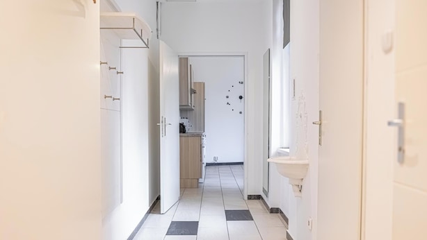 Un couloir étroit dot é d’un sol à carreaux, de murs blancs et d’une porte donnant sur un espace cuisine.
