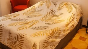 Sängyssä on lehtikuviollinen päiväpeite, siinä on punainen tuoli ja lattialla kuusikulmaiset laatat.