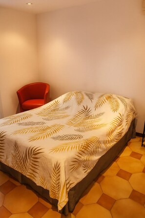 Ein Bett mit einem blattgemusterten Überwurf, ein roter Sessel und sechseckige Fliesen.