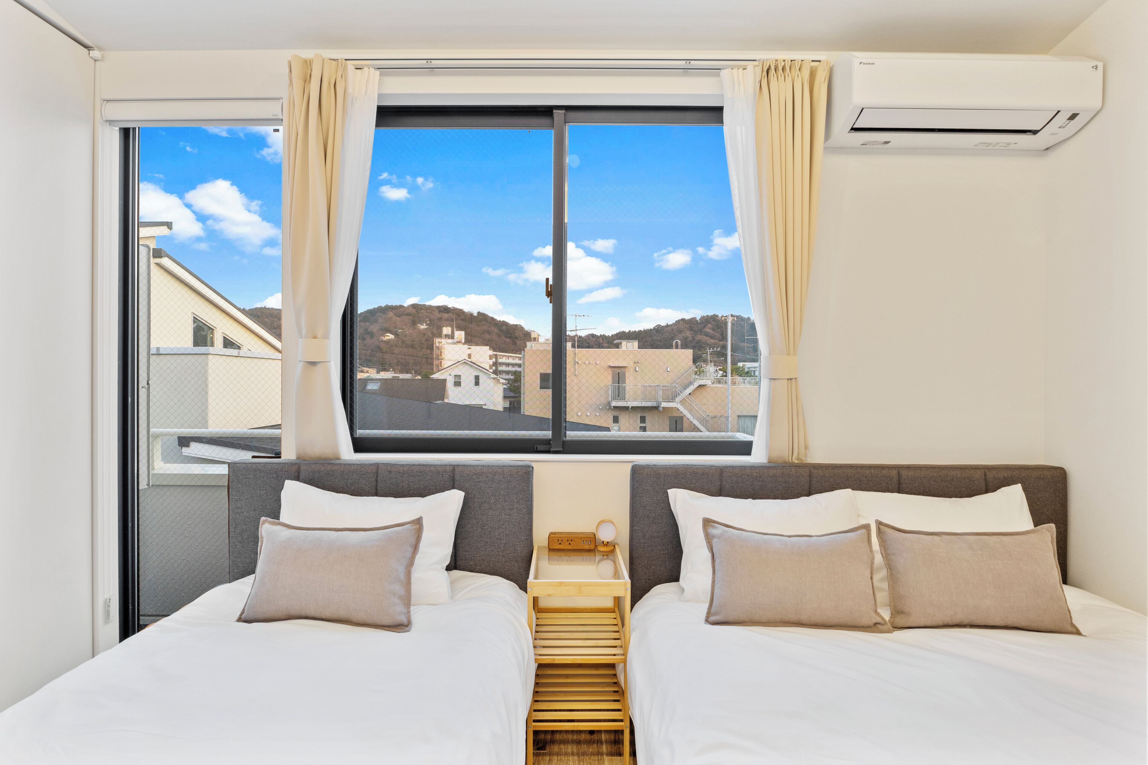 A hotel room with two beds, a window with curtains, an air conditioning unit, and a view of buildings and hills.