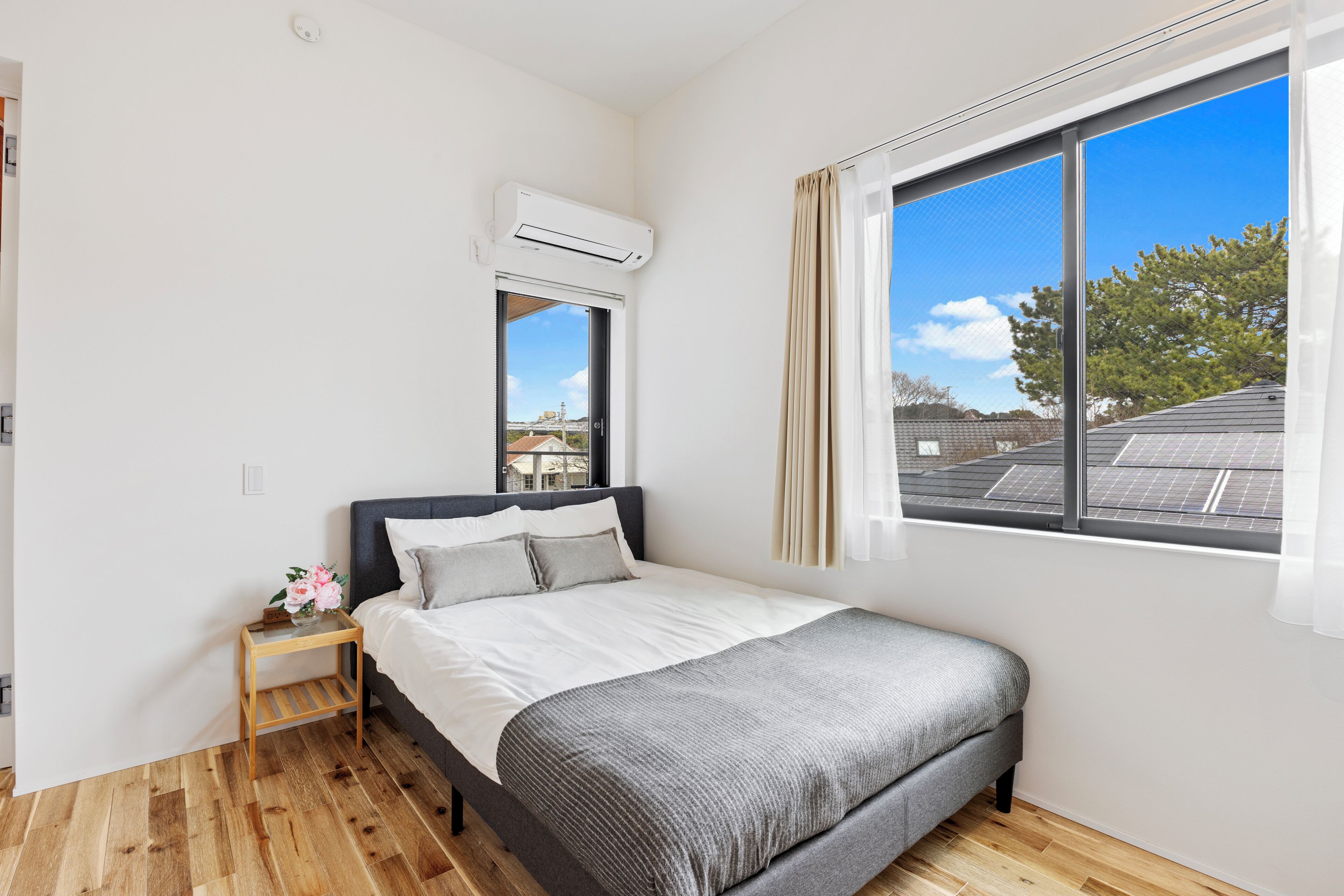Une chambre à coucher avec un lit, une fenêtre avec des rideaux, une petite table avec un vase et un appareil de climatisation.