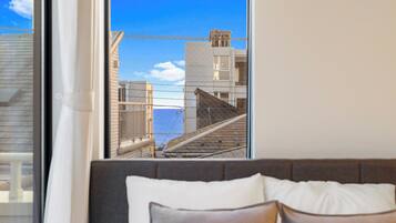 A bedroom with a large window overlooking a cityscape, a bed with a headboard, and a view of buildings and a body of water.