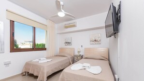 A hotel room with two beds, a ceiling fan, an air conditioner, a TV mounted on the wall, and a window with a view of buildings and trees. - Nissi Apartment #101 (Ayia Napa)