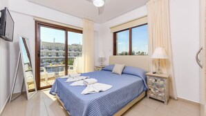 A hotel room with a bed, a nightstand, a ceiling fan, a TV, a balcony with a table and chairs, and a view of buildings outside. - Nissi Apartment #101 (Ayia Napa)