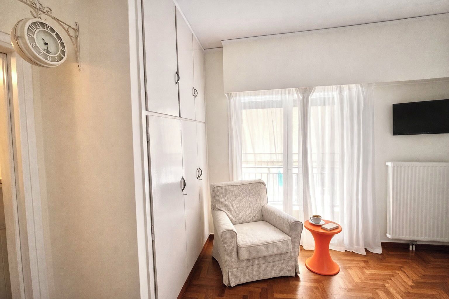 A room with a white wardrobe, a grey armchair, a small orange table, and a TV mounted on the wall.