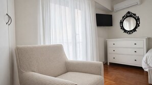 Una habitación con un sillón color beige, un aparador blanco, un televisor de pantalla plana montado en la pared y un espejo grande con un marco decorado.