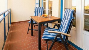 Ein Balkon mit Holztisch und zwei gestreiften Stühlen, gefliestem Boden und einem blau gerahmten Fenster.