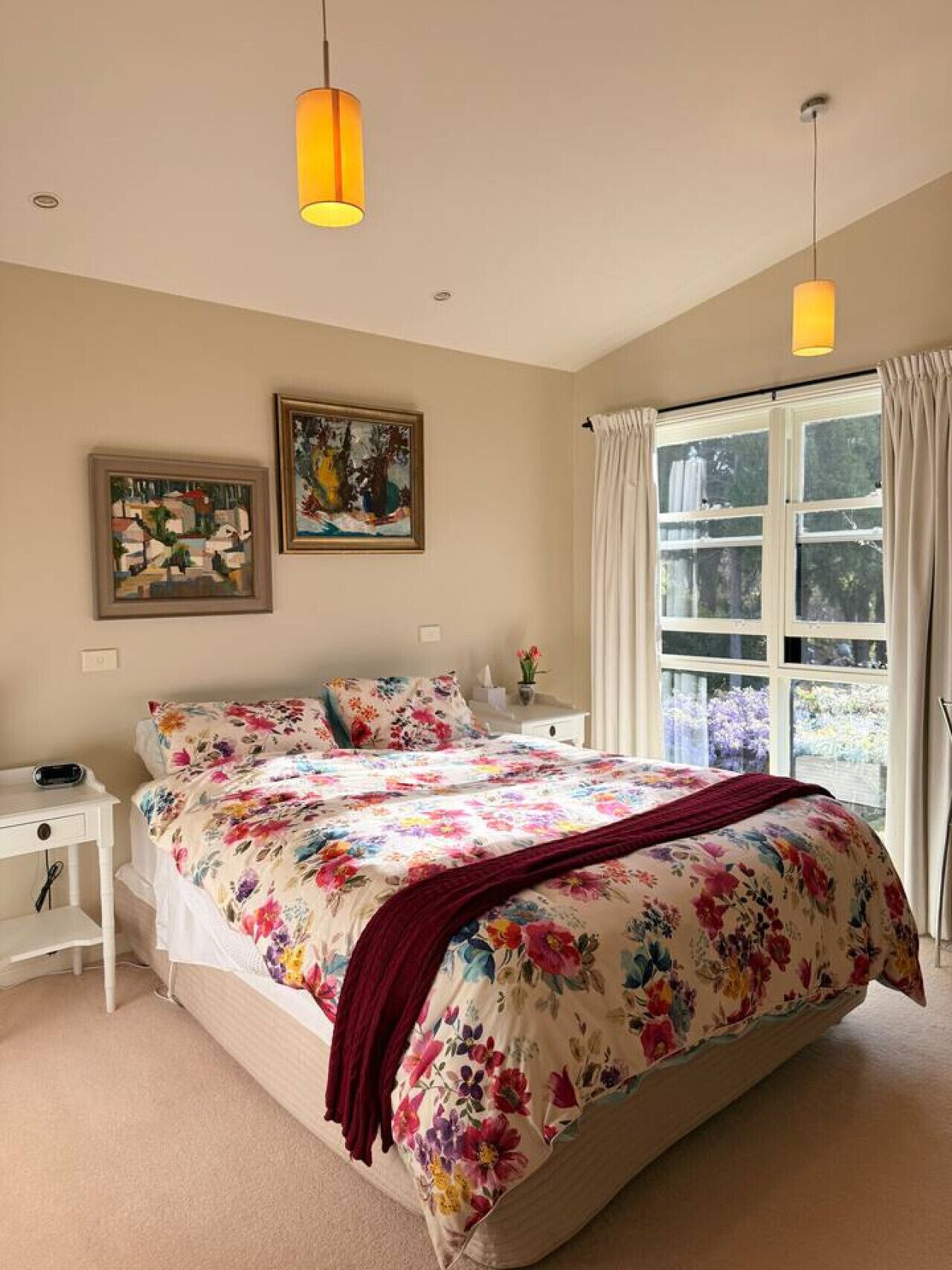 A bedroom with a large bed, two bedside tables, a nightstand, a window with curtains, and two hanging lamps.