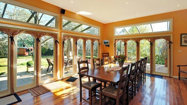 A spacious dining area with a large wooden table and multiple chairs, surrounded by large windows offering views of the outdoors. - Brecon Cottage | Serene 3BR Blue Mountains Escape (Leura)