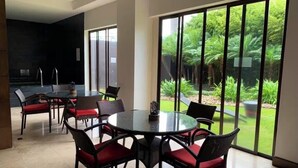 Un comedor moderno con una mesa redonda de cristal y sillas de mimbre negro con cojines rojos, grandes puertas correderas de cristal que dan a un jardín exuberante.
