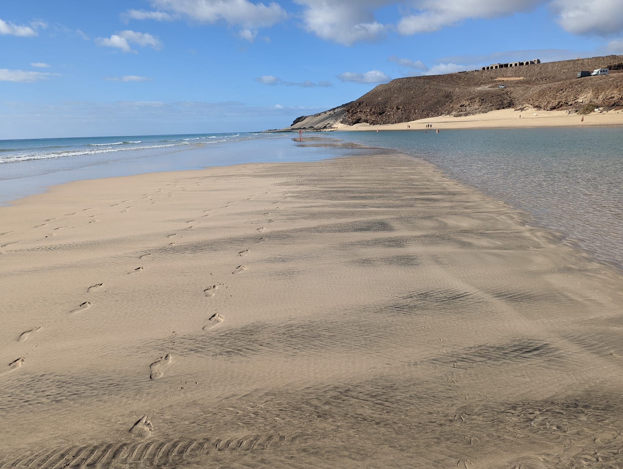 A sandy beach with footprints leading to the water, a clear sky, and a rocky hill in the background.