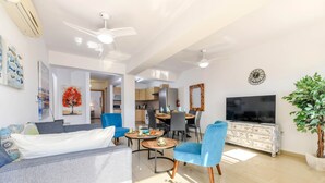 A modern living room with a sofa, chairs, a dining table, and a TV. There is a kitchen in the background with cabinets and a refrigerator. - Nissi Apartment 101 (Ayia Napa)