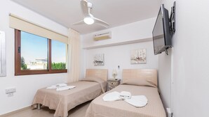 A hotel room with two beds, a ceiling fan, an air conditioner, a TV mounted on the wall, and a window with a view of buildings and trees. - Nissi Apartment 101 (Ayia Napa)