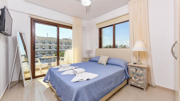 A hotel room with a bed, a nightstand, a TV, a balcony, and a view of buildings. - Nissi Apartment 101 (Ayia Napa)