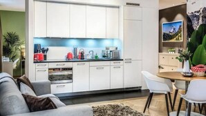 Een moderne keuken met witte kasten, een eethoek met een ronde tafel en witte stoelen, en een woonkamer met een grijze bank.