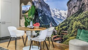 Een eetgedeelte met een houten tafel en witte stoelen, een grote plant en een landschapsmuur met een bergachtig panorama.