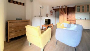 A living room with a wooden dresser, a yellow armchair, a blue armchair, a television on a wooden table, and a kitchen area with wooden cabinets and a staircase. - Brenlo (La Salle)
