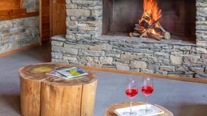 Ένα ζεστό δωμάτιο με ένα πέτρινο τζάκι, ξύλινα τοιχώματα και ένα στρογγυλό ξύλινο τραπέζι με δύο ποτήρια κόκκινο κρασί.