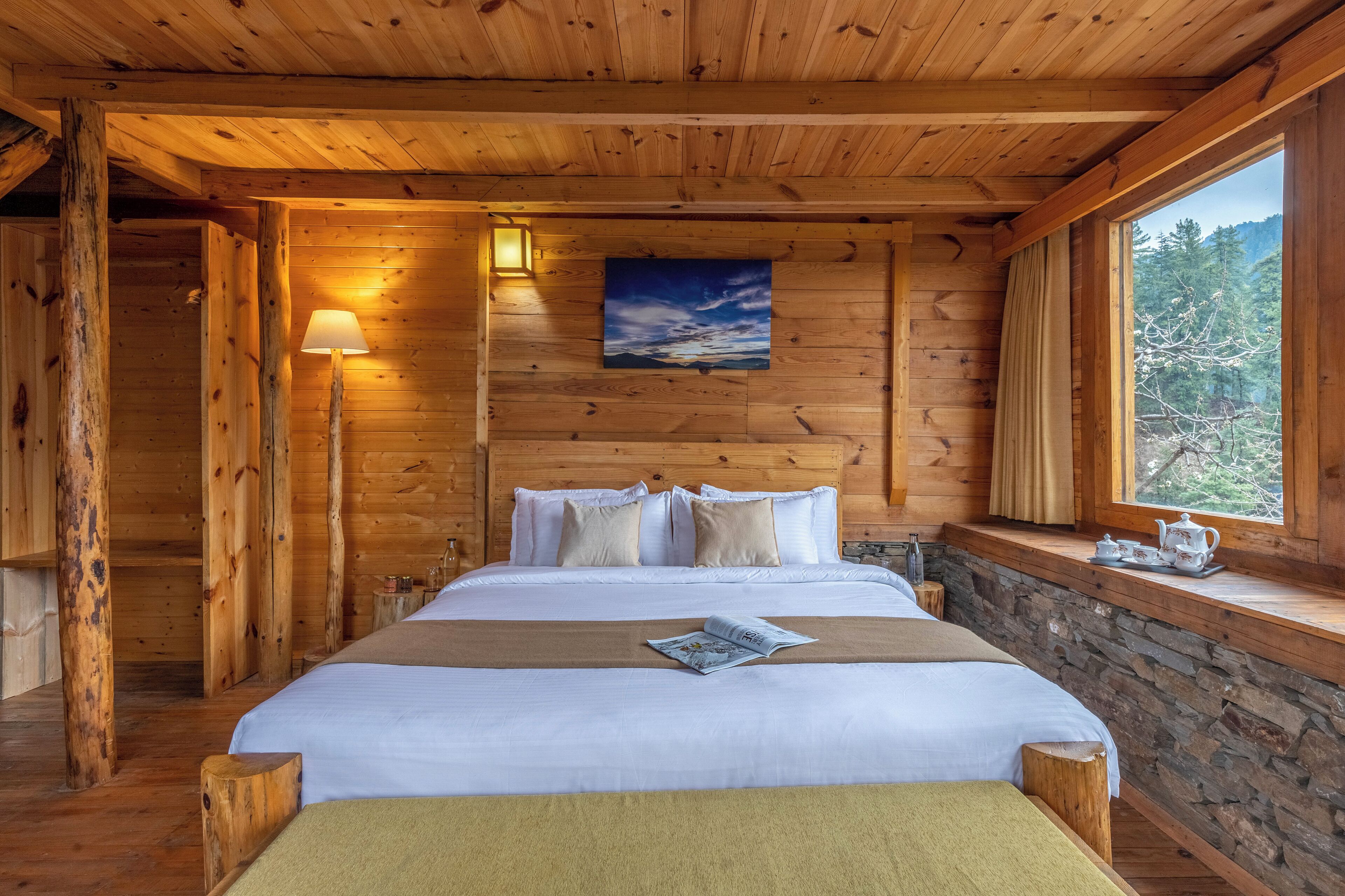 A wooden cabin bedroom with a bed, a nightstand, a window with a view of trees, and a painting on the wall.