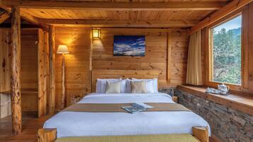 A wooden cabin bedroom with a bed, a nightstand, a window with a view of trees, and a painting on the wall.