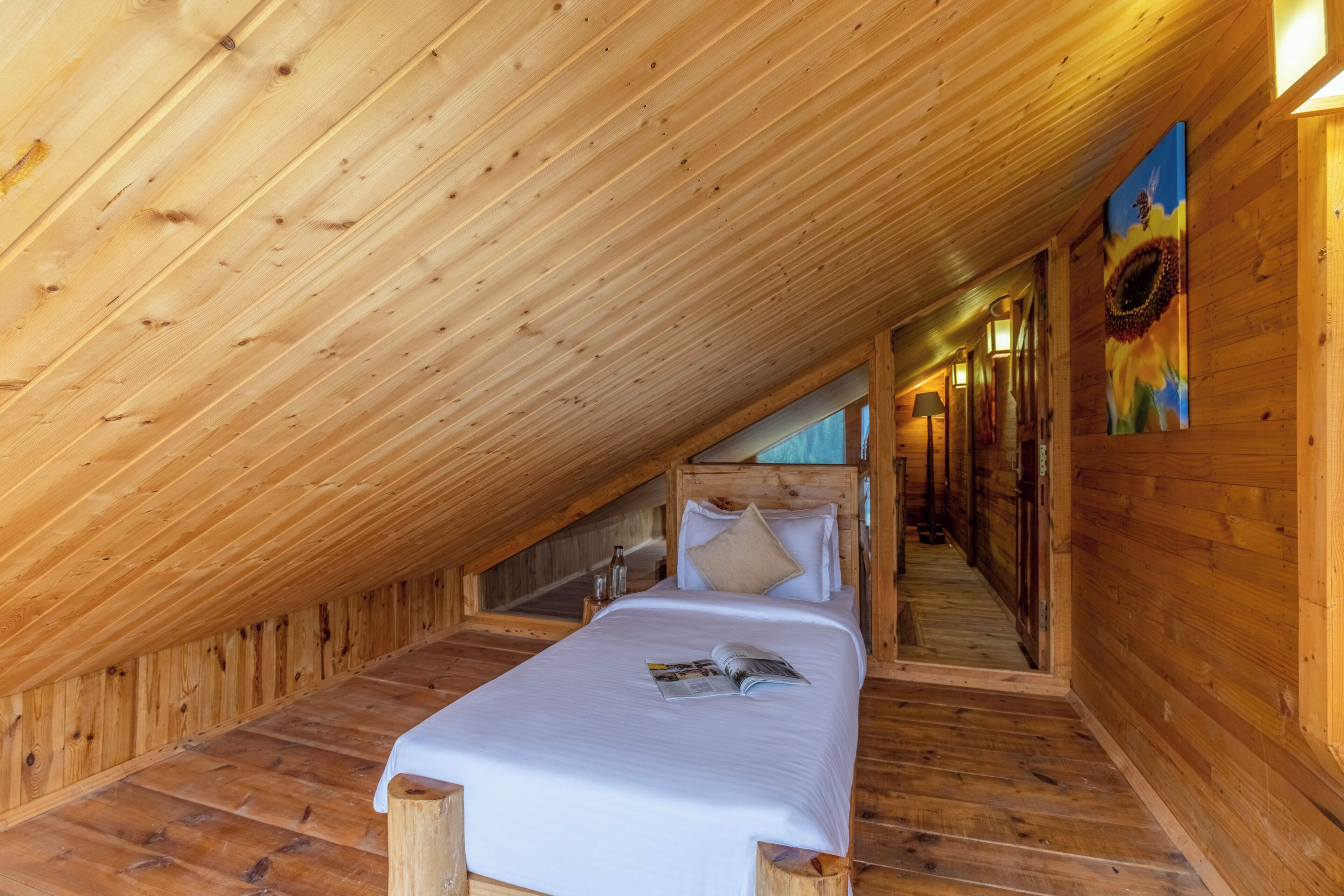 A wooden interior with a sloped ceiling, a bed with white bedding, and a painting of a sunflower on the wall.