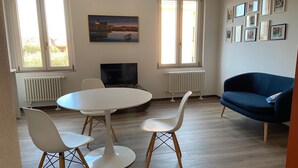 Ein Wohnzimmer mit rundem Tisch, zwei weißen Stühlen, einem blauen Sofa, einem Fernseher und einem Bild an der Wand.