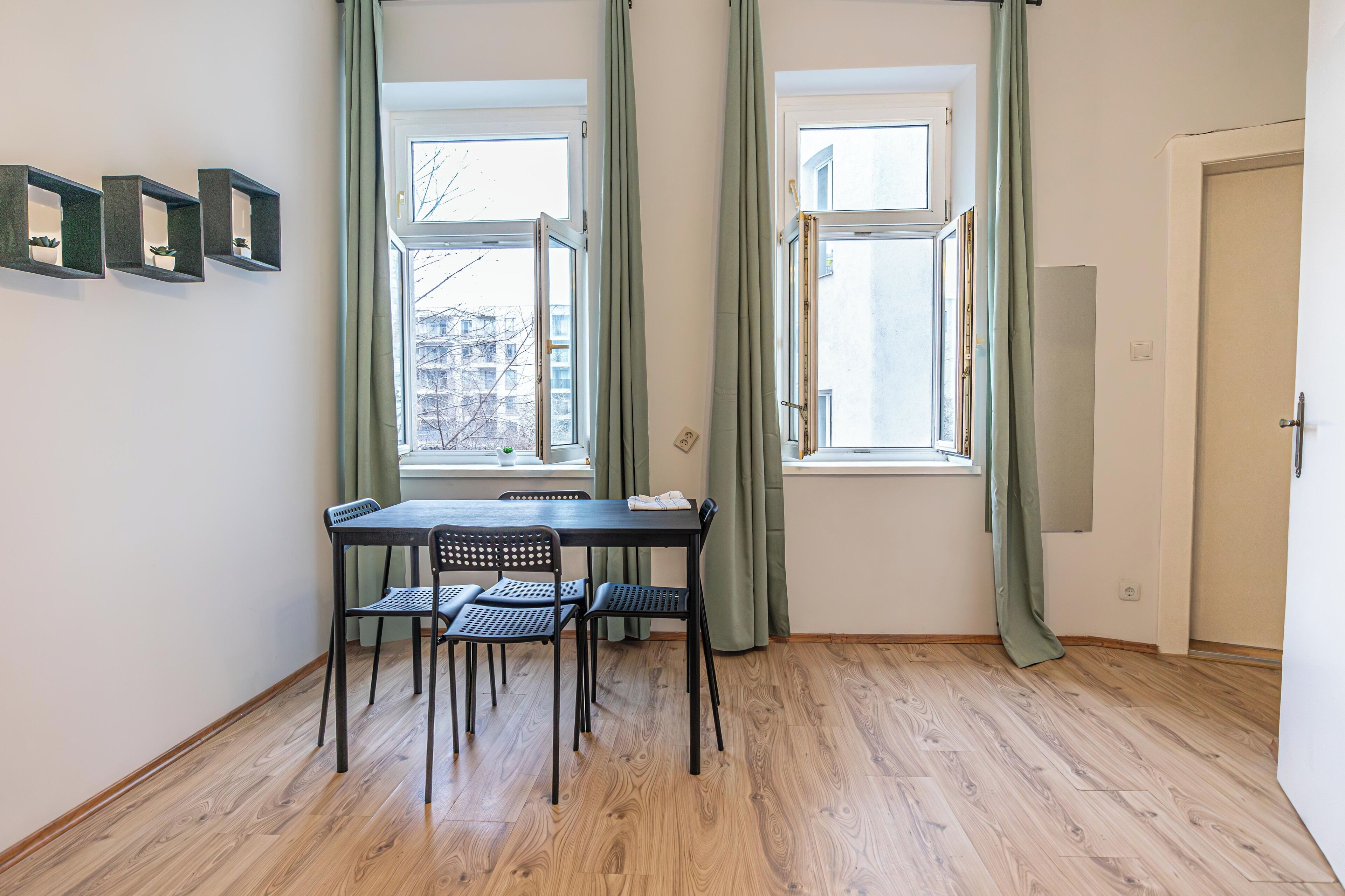 A room with a wooden floor, a table with four chairs, two windows with curtains, and a door.