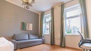 Ett modernt vardagsrum med en grå soffa, ett trägolv, en kristallkrona och en utsikt genom ett fönster med gardiner.