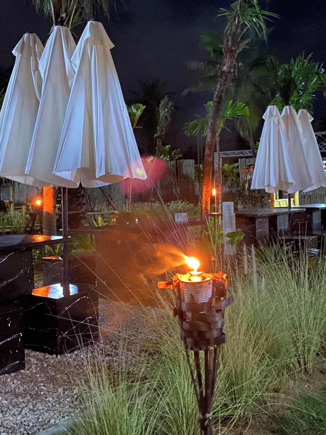 Mobiliario exterior con sombrillas blancas, una chimenea pequeña y palmeras al fondo.